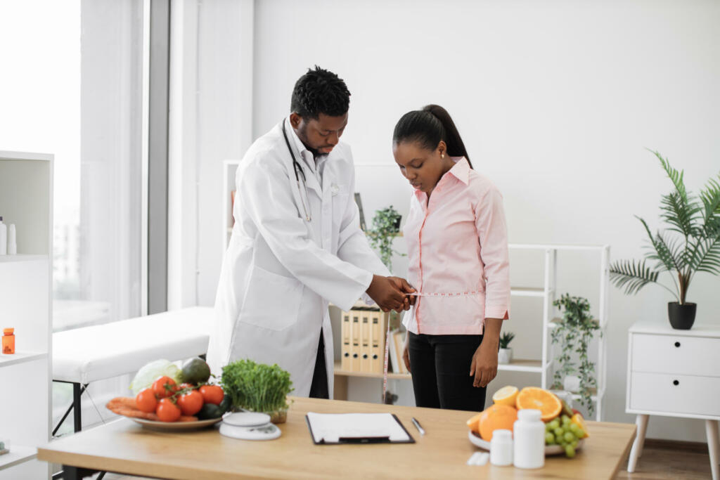 Multiethnic man in doctor's coat finding out waist length of female patient with measuring tape in consulting room. Efficient dietitian calculating body mass index for assessing healthy weight.