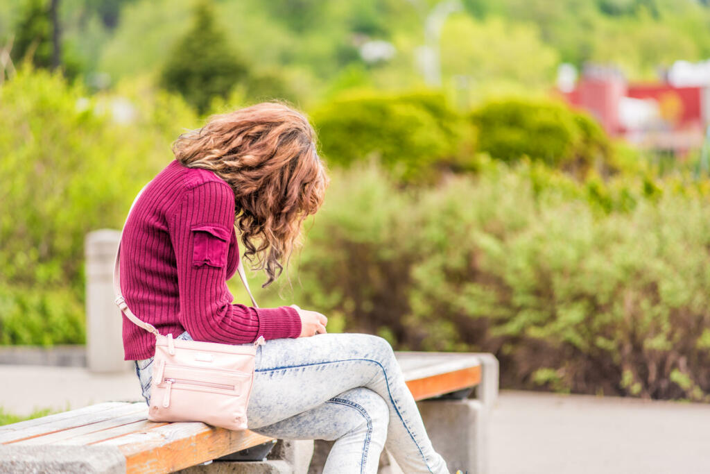 One young happy woman sitting on bench on sidewalk in green downtown city park in Saguenay, Canada, Quebec during summer looking at phone or reading book