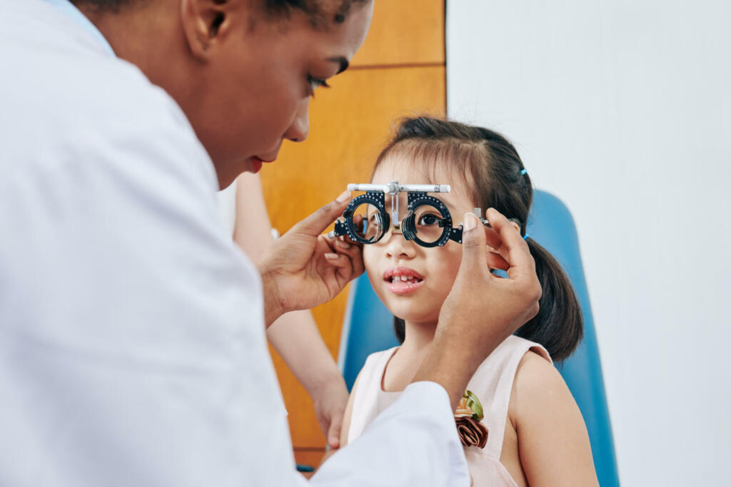 Pediatric optometrist helping little girl to put on test glasses when checking her eyesight