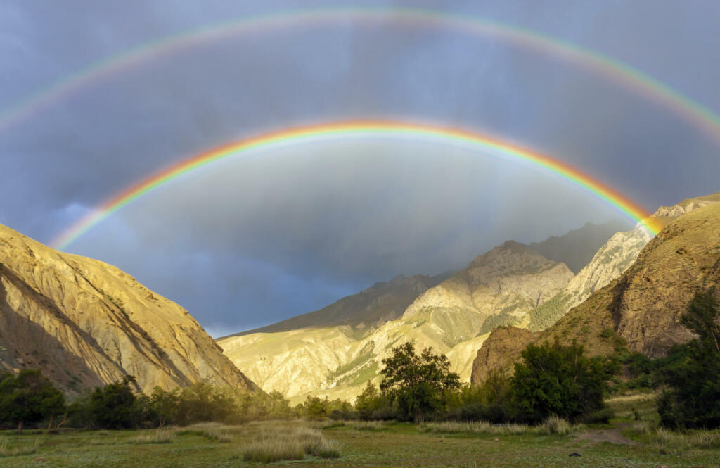 Rare double rainbow in the dark sky in the mountains. Double multicolored arc and band of Alexander between them before thunderstorm in a mountain valley. Atmospheric, optical and meteorological phenomenon observed when the sun illuminates water droplets of rain, composed of colors of the visible radiation spectrum.