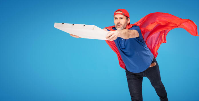 Super hero pizza delivery guy flying on blue studio background, wearing red cape.  A lot of copy space.