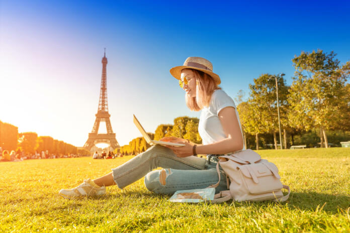 An Asian girl works on a laptop sitting on the grass on the Champ de Mars in Paris overlooking the Eiffel tower at sunset. Concept of remote work and freelancing