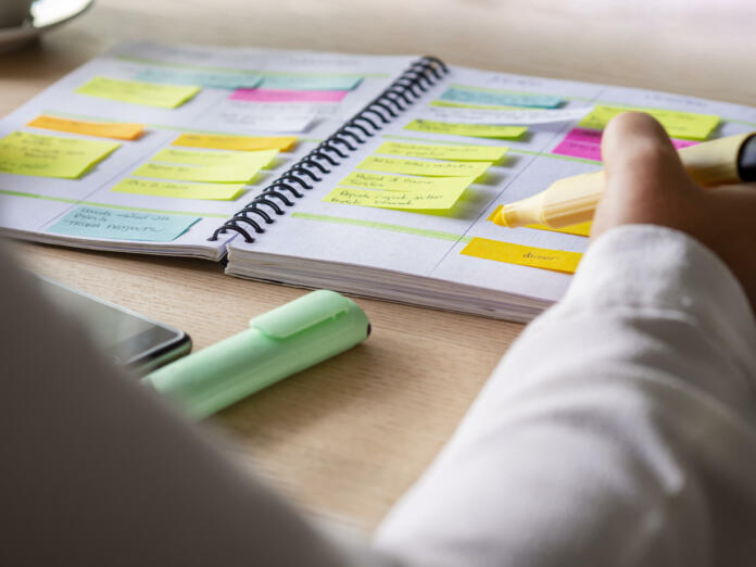 Close-up of agenda organize with color-coding sticky notes for time management. Productive schedule for appointments and reminders. Hand holding a yellow highlighter marker. Organization and planning