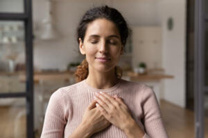 Close up of calm young Caucasian woman hold hands on heart chest feel grateful and thankful. Happy millennial female show gratitude, love and care, pray or visualize. Religion, faith concept.