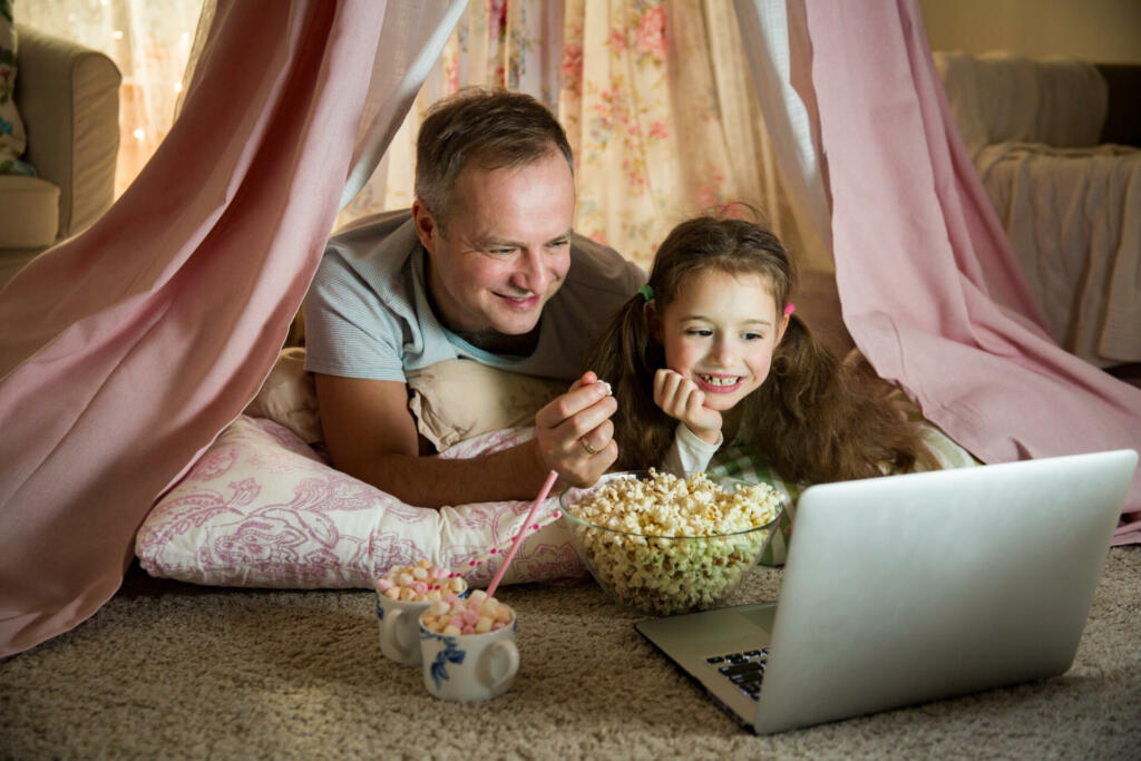 Family quality time. Father and daughter lie in homemade pink tent with flowers, watch cartoons on laptop, eating popcorn, laugh. Cozy stylish room. Family bonds concept
