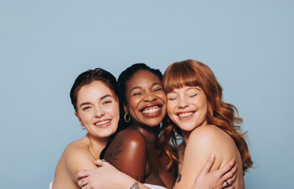 Group of happy women with different skin tones smiling and embracing each other. Three diverse women feeling comfortable in their natural skin. Body positive young women standing together in a studio.