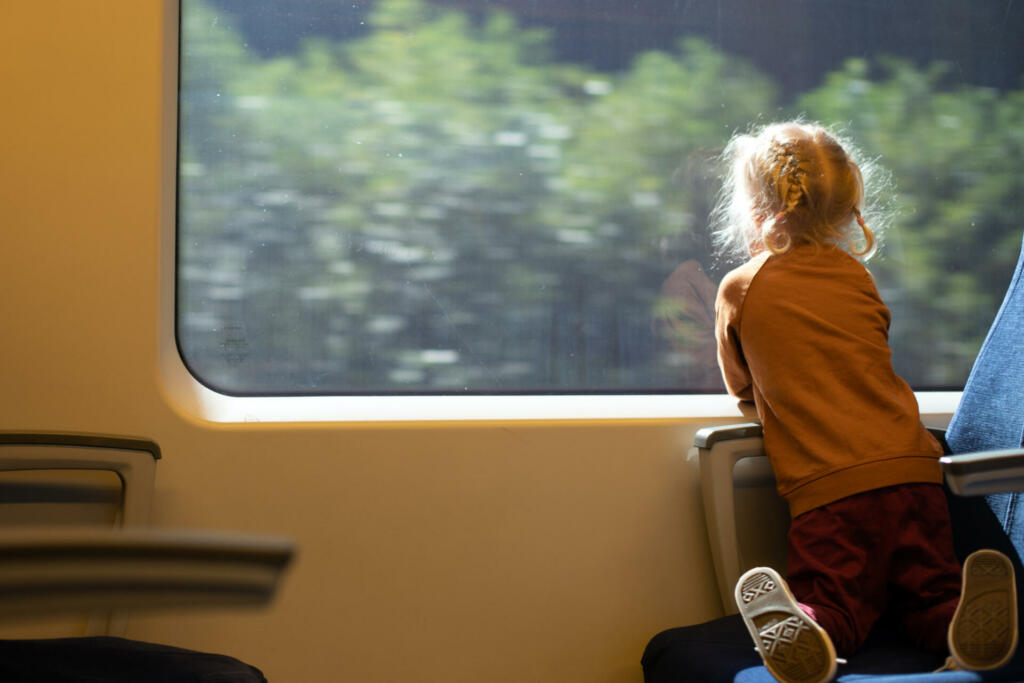 Little girl 3-4 years with blonde hair watching window while travelling by train. Public transportation, kid back view