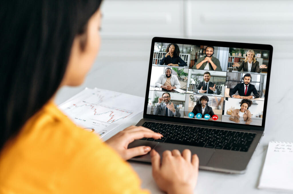 Online video conference, group brainstorm. Over shoulder view of girl on a laptop screen with group of multiracial business people talking on a video call, discussing business project, business plan