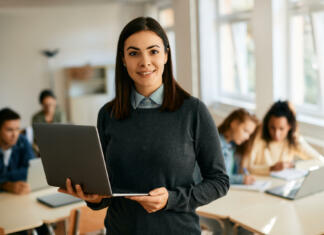 Teh 5 stvari študent ne sme nikoli pozabiti Smiling teacher using laptop during computer class at high school and looking at camera. Her students are learning in the background.