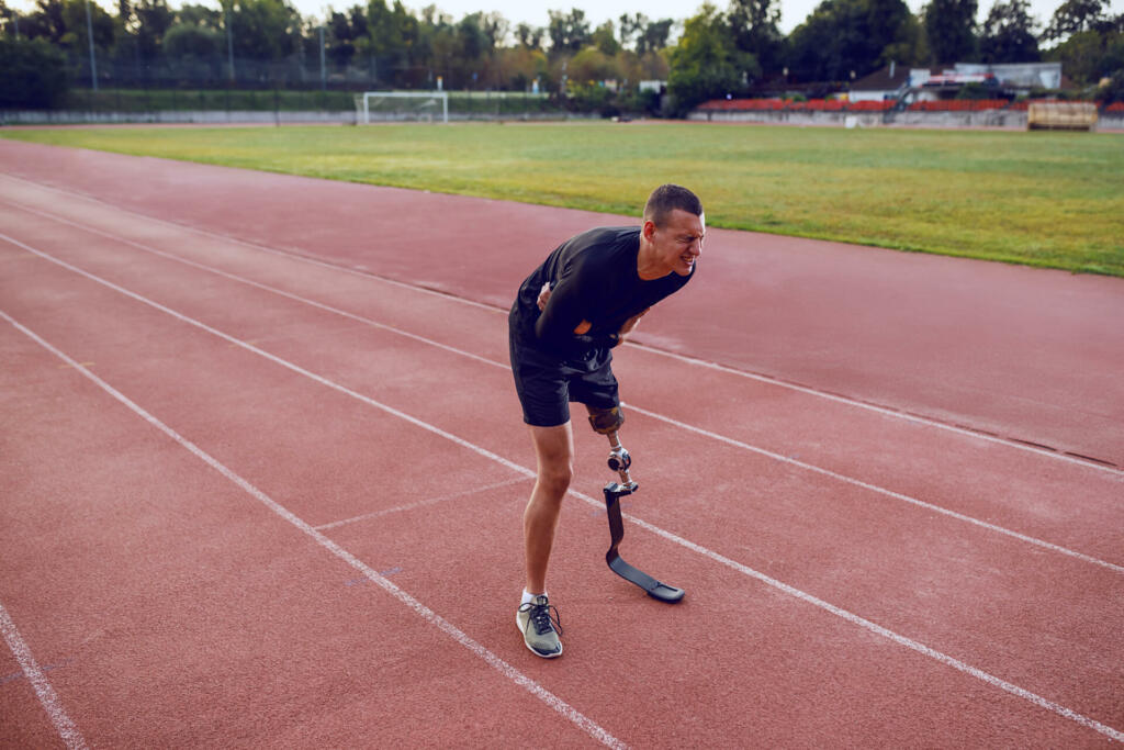 Sporty caucasian man with artificial leg standing on running track and having pain in stomach.