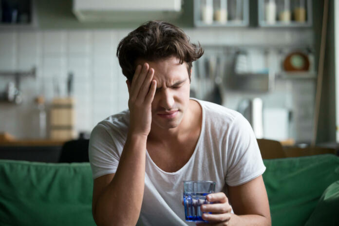 Young man suffering from strong headache or migraine sitting with glass of water in the kitchen, millennial guy feeling intoxication and pain touching aching head, morning after hangover concept