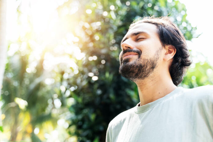 A bearded man is meditating outdoor in the park with face raised up to sky and eyes closed on sunny summer day. Concept of meditation, dreaming, wellbeing and healthy lifestyle