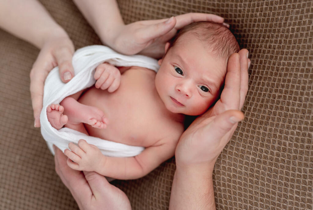 Awake naked newborn framed by parents hands, top view