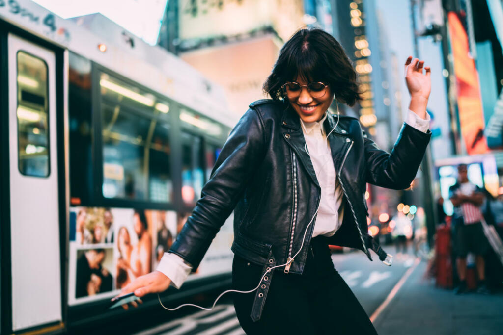 Carefree woman in fashionable clothing listening funny audio and dancing during evening walk in New York City, cheerful female enjoying adolescent music podcast during nightlife lifestyle in Manhattan