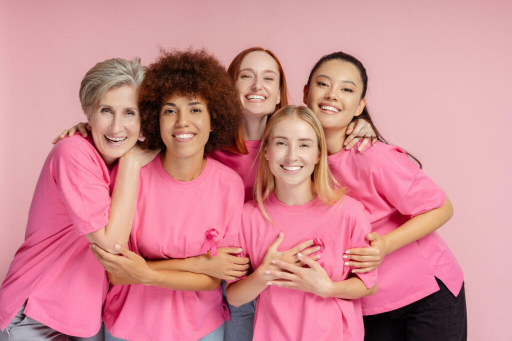 Group of smiling women wearing t shirts with pink ribbon isolated on pink background. Breast cancer