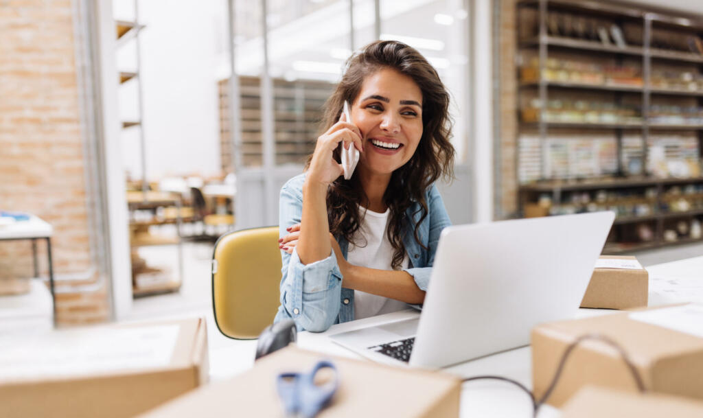 Happy young businesswoman speaking on the phone while working in a warehouse. Online store owner making plans for product shipping. Creative female entrepreneur running an e-commerce small business.
