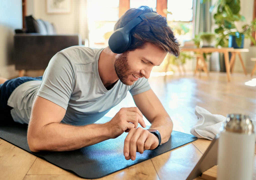One fit young caucasian man listening to music with headphones and checking time on digital wristwatch while exercising at home. Guy wearing fitness tracker on arm to monitor progress, heart rate and calories burned during training workout
