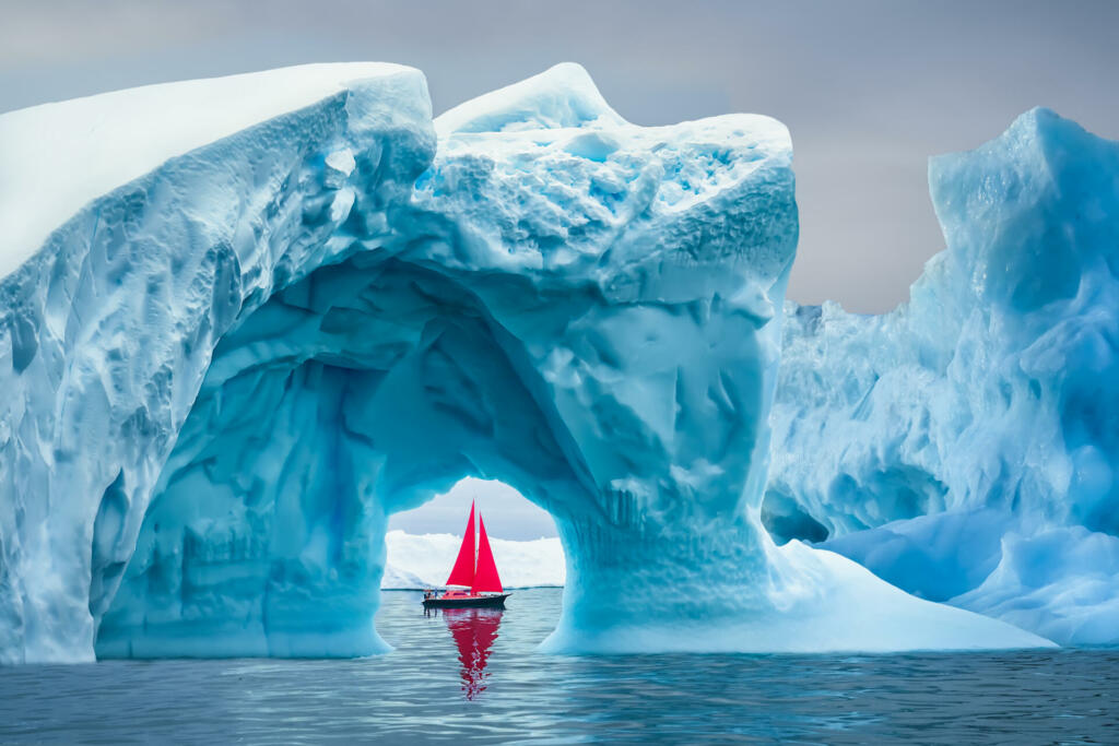 Red sailboat sailing under a majestic iceberg arch on sunny blue Artic Ocean in Greenland. Sailing through enormously huge icebergs near Ilulissat Icefjord. Sail boat with red sails cruising among icebergs during midnight sun season. Ship sailing past majestic icebergs on sunny blue Artic Ocean, Ilulissat Icefjord, Ilulissat, Disko Bay, Greenland, Unesco World Heritage Site