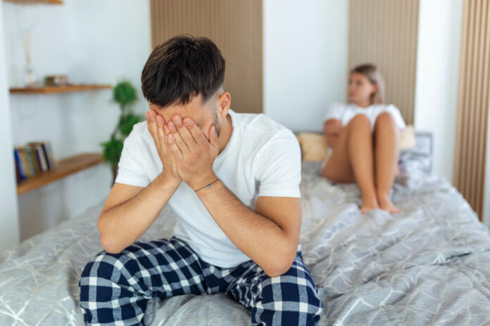 Sad thoughtful man after arguing with girlfriend.Relationship difficulties, conflict and family concept,unhappy couple having problems at bedroom. Sad guy sitting on bed,girlfriend in the background