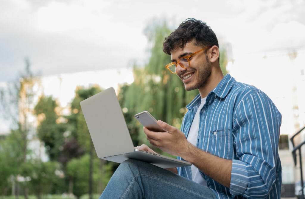 Young handsome Indian man using laptop computer, mobile phone, working freelance project online, sitting outdoors. Successful business. Asian student studying, learning language, online education concept