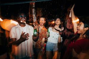 A cheerful group of multiracial friends is celebrating and having fun with confetti at the open-air summer rooftop nightclub. They are having a blast, dancing and drinking beer.