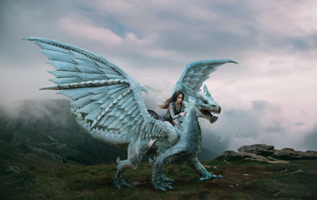 A young woman sits astride a dragon. Fantasy photography. A huge creature with spikes and wings stands on top of a mountain. Beautiful nature sky and white clouds