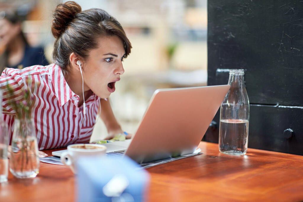 astonished young female watching content on her laptop with headphones in ears, can't believe what she saw, with her mouth open. amazed, astonished reaction concept