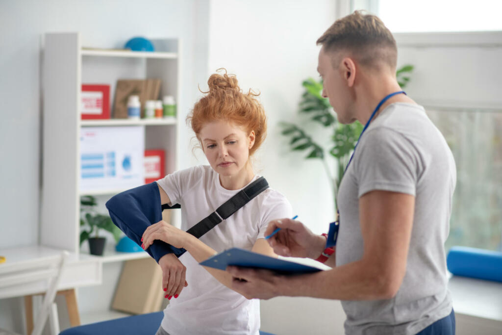 At rehabilitation center. Male physiotherapist writing prescriptions, female patient touching her arm sling