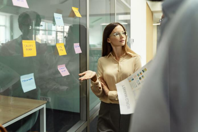 Businesswoman wearing stylish eyeglasses using sticky notes, agile methodology for productivity working together in modern office. Scrum master planning strategy, talking with business colleagues