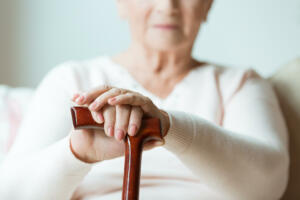 Elder holds hands on walking stick while sitting on white sofa in nursing home
