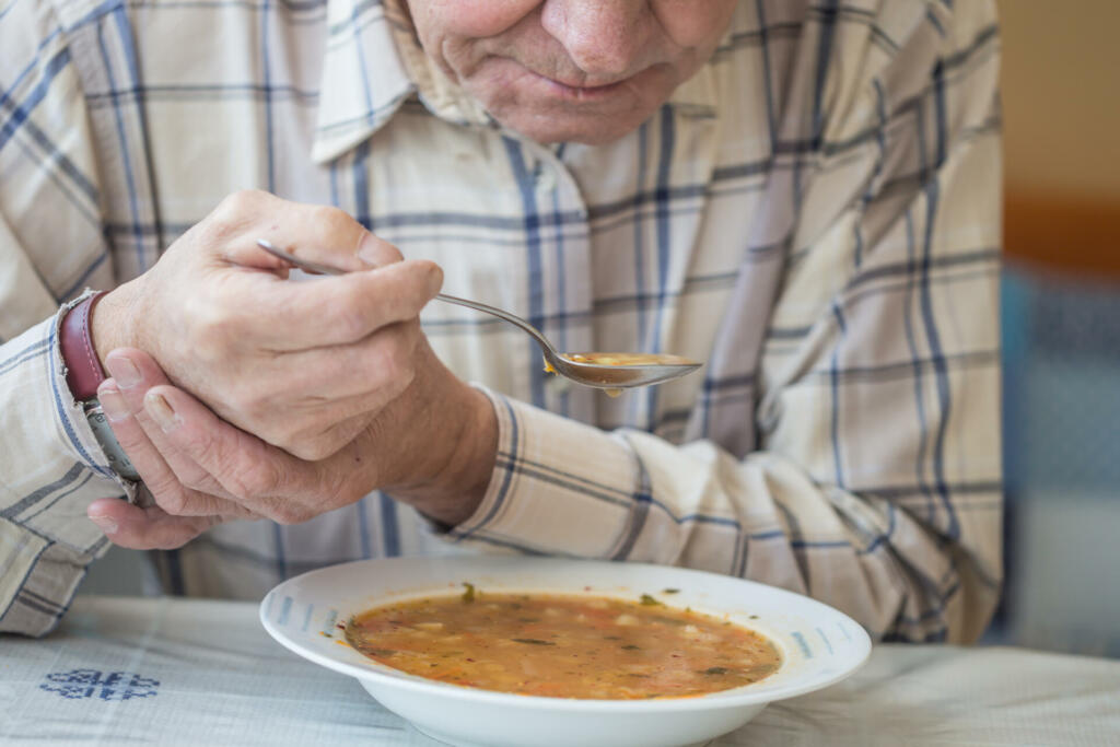 Elderly man with Parkinsons disease holds spoon in both hands.