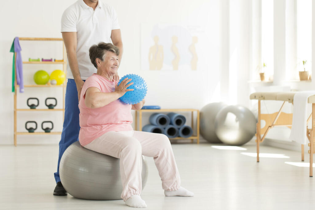 Happy senior woman sitting on grey ball and holding blue ball while exercising at gym