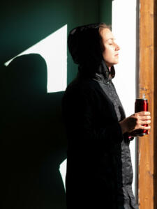 Happy young woman in black raincoat against wall basking in sun autumn day. Smiling girl drinking tea with thermo can bottle. Lifestyle real people Seasonal depression mental health, sunrays Vitamin D