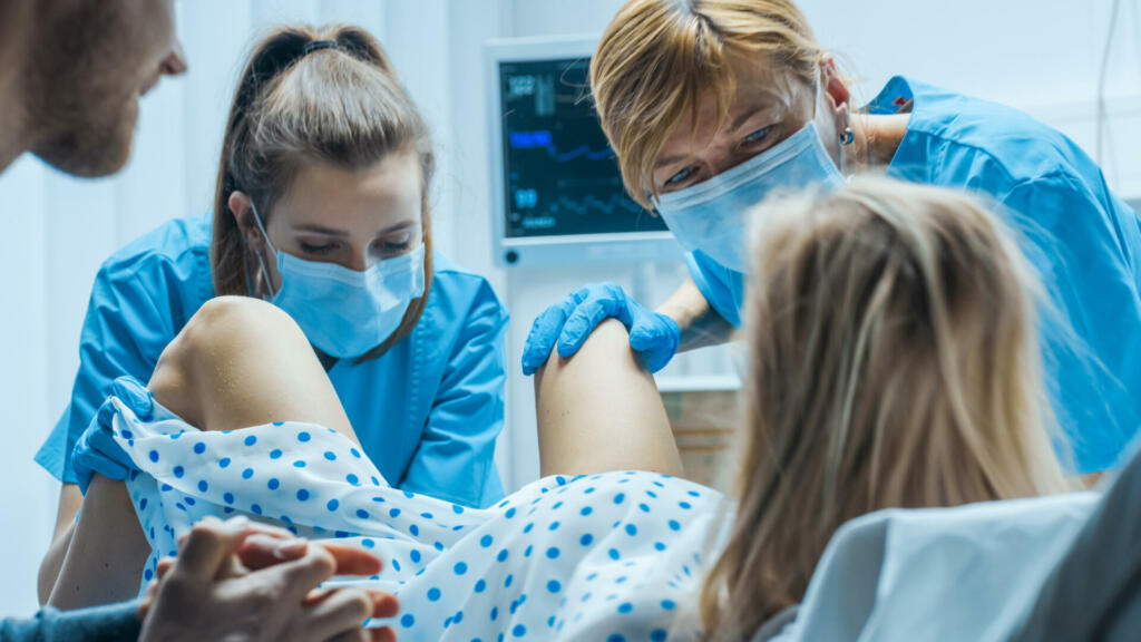 In the Hospital Woman in Labor Pushes to Give Birth, Obstetricians Assisting, Husband Holds Her Hand. Modern Delivery Ward with Professional Midwives. Back View Footage.