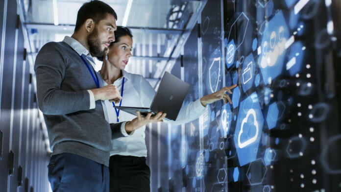 Male IT Specialist Holds Laptop and Discusses Work with Female Server Technician. They're Standing in Data Center, Rack Server Cabinet with Cloud Server Icon and Visualization.