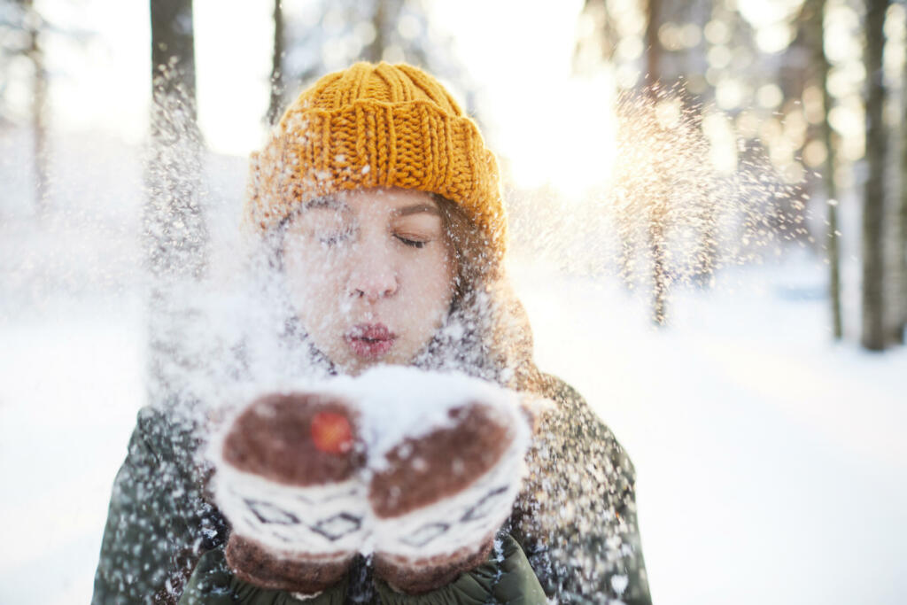 Portrait of happy young woman playing with snow in winter, blowing snowflakes to camera, copy space