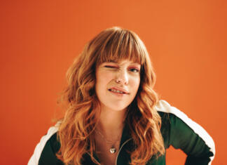 Ali rdečelasci ne čutijo bolečine in so rdečelaske so bolj odporne? Quirky young woman winking at the camera while standing in a studio. Young woman with ginger hair standing against a vibrant orange background in casual clothing.