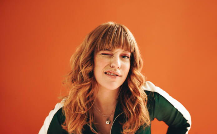 Quirky young woman winking at the camera while standing in a studio. Young woman with ginger hair standing against a vibrant orange background in casual clothing.