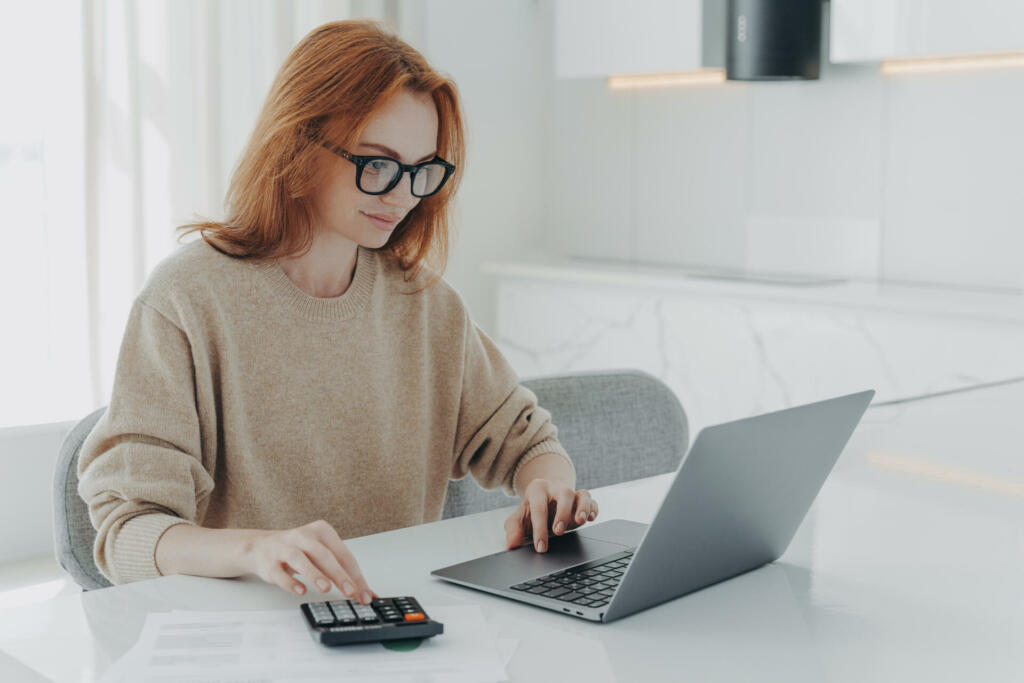 Redhead European female accountant analyzes documents uses calculator makes report uses laptop computer sits at white desktop wears spectacles beige jumper poses against white home interior.