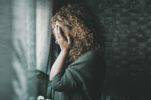 Sad and worried woman side portrait at home in the dark looking outside the window. Worried and lonely female alone indoor. Concept of end of relationship and unsuccessful emotion. Lady touching face