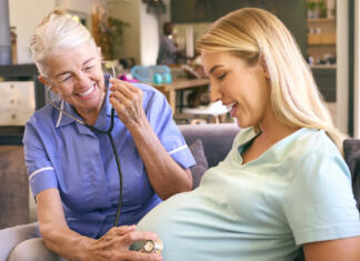Babištvo kmalu na seznamu Unesca? Senior Midwife With Stethoscope Visiting Pregnant Woman At Home With Family In Background