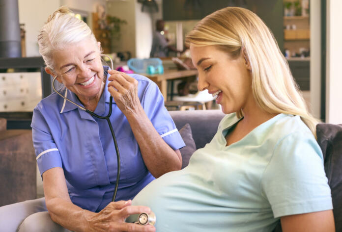Senior Midwife With Stethoscope Visiting Pregnant Woman At Home With Family In Background
