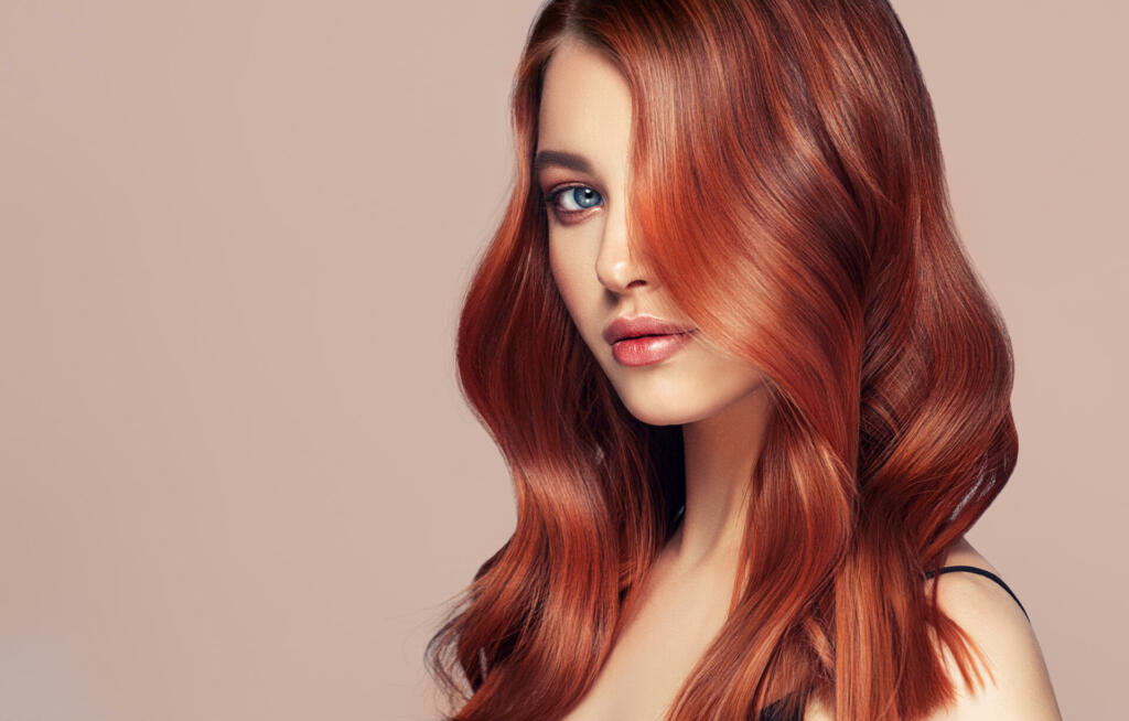 Young, red haired woman with soft, kind and tender look of blue eyes at viewers. Beautiful model with long, curly hairstyle and vivid makeup. Perfect dense, wavy,and shiny hair. Hairdressing art, hair care and beauty products.