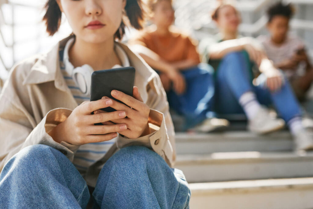 Closeup of teenage girl holding smartphone outdoors while sitting on metal stairs with group of friends in background, copy space