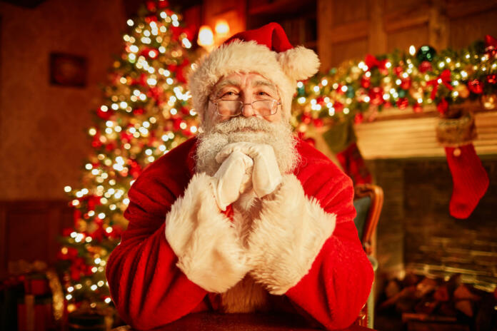 Front view portrait of traditional Santa Claus smiling at camera resting head on arms with christmas tree in background