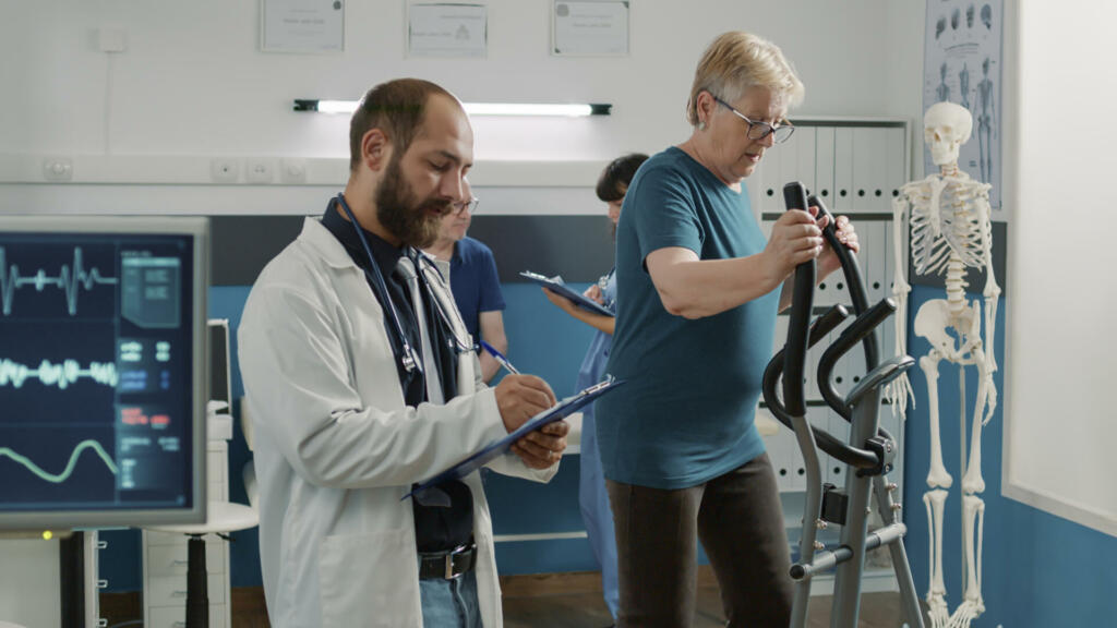 Osteopath taking notes and watching patient use stationary bicycle, doing physical exercise for muscle recovery. Treat mechanical disorders with electrical bike and heart rate monitor.