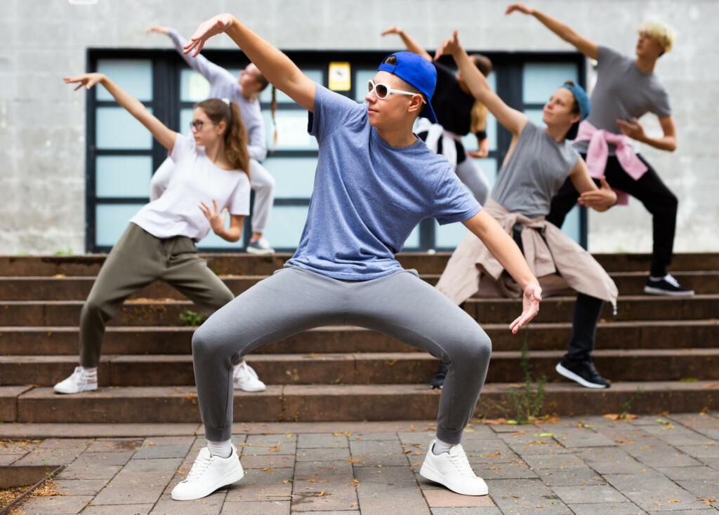 Positive teen boy dancing modern street dance outdoors with teenagers in background
