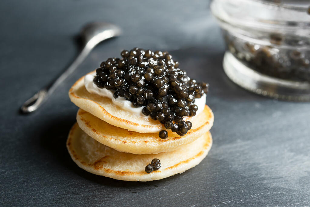 Black sturgeon caviar on small blini pancakes, a glass jar and a spoon on a dark gray background