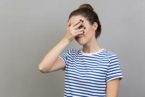 Portrait of interested woman wearing striped T-shirt covering eyes with hand, peeking through fingers with happy curious expression. Indoor studio shot isolated on gray background.