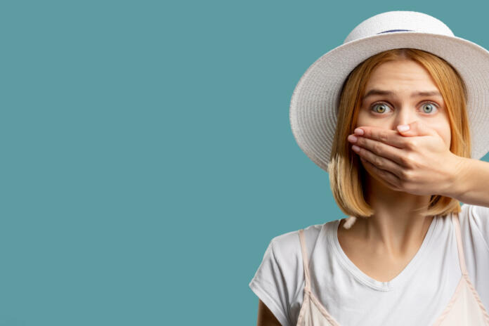 Speech freedom. Female rights. Scared woman in white closing mouth hand looking at camera isolated on blue copy space. Keep secret. Advertising background